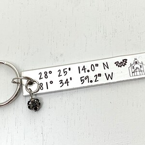 May include: A silver-toned rectangular metal keychain engraved with the coordinates 28° 25' 14.0" N and 81° 34' 59.2" W, featuring a small etched bat and a building icon, accompanied by a small round charm with a dark, textured surface.