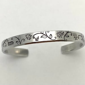 May include: A silver bracelet with an engraved design of vines, leaves, and ladybugs. The design is in black and the bracelet is polished.