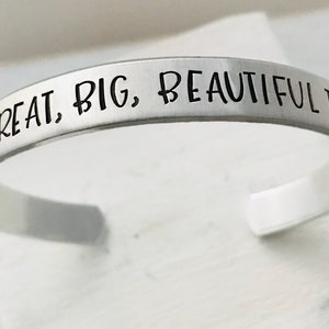 May include: A silver-colored metal cuff bracelet with a smooth, brushed finish. The outer surface is engraved with the black text "IT'S A GREAT, BIG, BEAUTIFUL TOMORROW". The bracelet features an open-ended design, allowing for adjustment to fit the wrist.