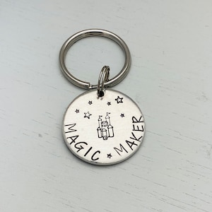 May include: A silver-colored keychain with a round charm. The charm has the words "MAGIC MAKER" around the edge, with a castle illustration in the center. Small star shapes are stamped around the castle. A silver key ring is attached.