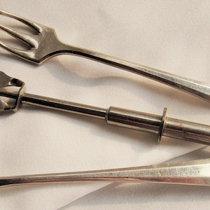 May include: Two silver-toned metal forks with three tines and a central metal tool featuring a two-pronged head, a cylindrical shaft, and a small circular plunger at the end. The forks have smooth, tapered handles with a small engraved monogram.