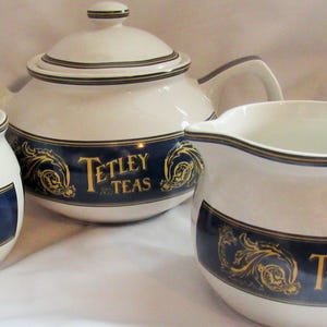 May include: A white ceramic tea set comprising a teapot, sugar bowl, and milk jug. Each item features a navy blue band with gold floral patterns and the text "Tetley Teas". The set has black and gold trim.