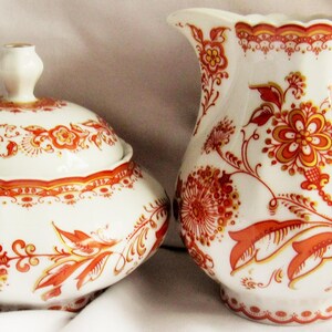 Puede incluir: Un azucarero de cerámica blanco con tapa y una jarra a juego. Ambas piezas están decoradas con un estampado floral en tonos naranja y rojo. El azucarero tiene forma redondeada y la jarra tiene un asa curvada.