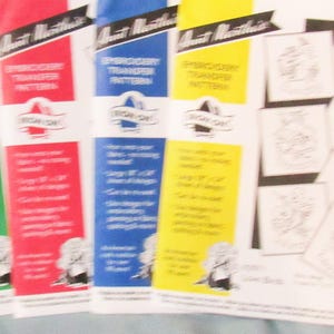 May include: Four Aunt Martha's embroidery transfer pattern packets in green, red, blue, and yellow. Each packet features the text Embroidery Transfer Pattern and Iron On, with a large 20.32 cm by 35.56 cm sheet of designs for embroidery or fabric painting.