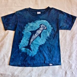 May include: A blue tie-dye t-shirt with a white hammerhead shark graphic. The shirt has a crew neck and short sleeves.