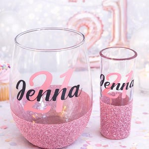 May include: A clear wine glass and shot glass with pink glitter bases. Both glasses have the number "21" and the name "Jenna" in pink and black lettering. The background includes a banner that says "Happy 21st".