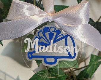 May include: A clear round ornament with a white ribbon. The ornament has a blue and silver megaphone with a white pom-pom and the name "Madison" written in white.