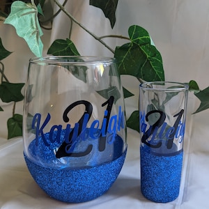 May include: Two personalized glasses with blue glitter bottoms. The larger glass is a wine glass with the name "Kayleigh 21" on it. The smaller glass is a shot glass with the text "21" on it.