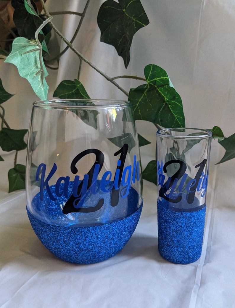 May include: Two personalized glasses with blue glitter bottoms. The larger glass is a wine glass with the name "Kayleigh 21" on it. The smaller glass is a shot glass with the text "21" on it.