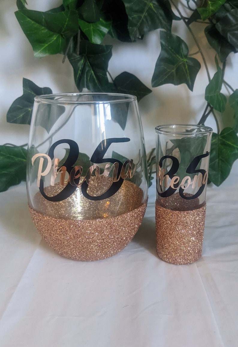 May include: Two gold glitter glasses with black vinyl lettering. The larger glass says "35 Pheona" and the smaller glass says "35 Pheona".