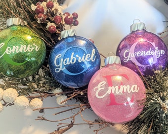 May include: Four personalized Christmas ornaments with glitter. The ornaments are green, blue, purple, and pink. The names on the ornaments are Connor, Gabriel, Gwendolyn, and Emma.