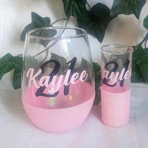 May include: Two pink glasses with glitter bottoms. The larger glass is a wine glass with the text "21 Kaylee" on the front. The smaller glass is a shot glass with the text "21 Kaylee" on the front.