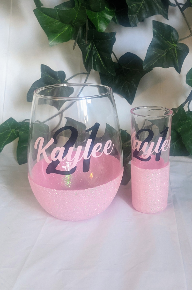 May include: Two pink glasses with glitter bottoms. The larger glass is a wine glass with the text "21 Kaylee" on the front. The smaller glass is a shot glass with the text "21 Kaylee" on the front.