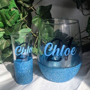 May include: Two blue glitter glasses with the name "Chloe" and the number "21" on them. The glasses are on a white surface.