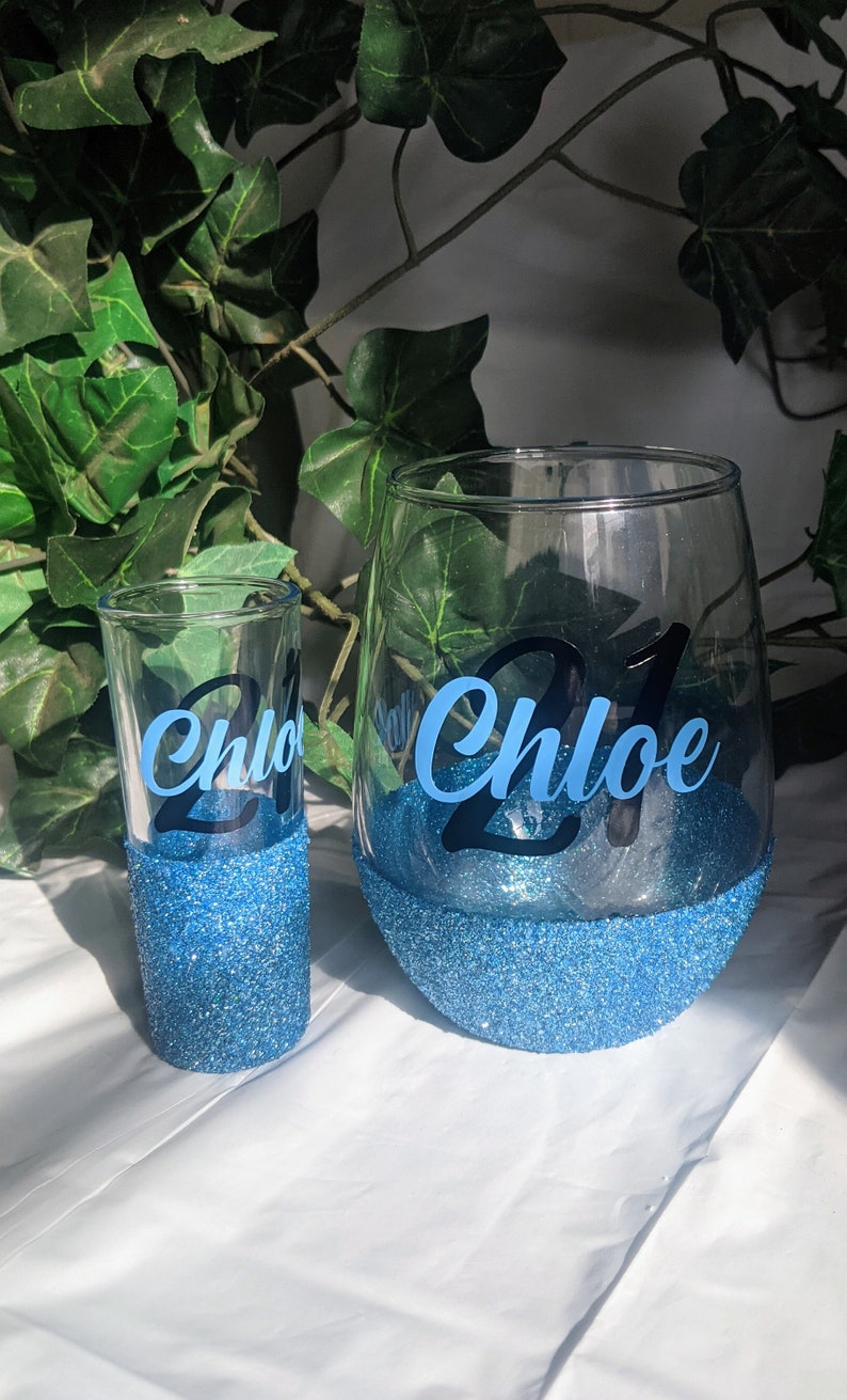 May include: Two blue glitter glasses with the name "Chloe" and the number "21" on them. The glasses are on a white surface.