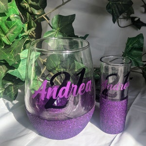 May include: Two purple glitter glasses with the name "Andrea" and the number "21" on them. The glasses are on a white surface.