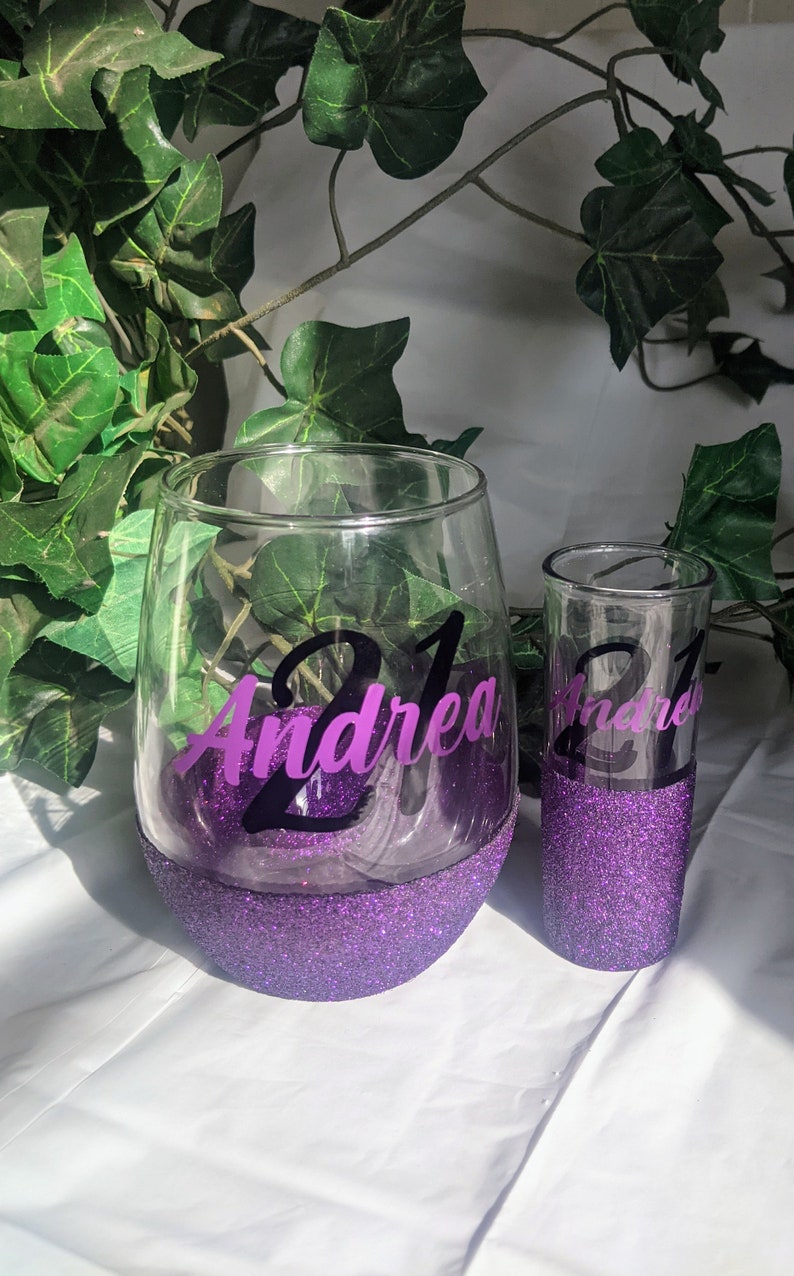 May include: Two purple glitter glasses with the name "Andrea" and the number "21" on them. The glasses are on a white surface.