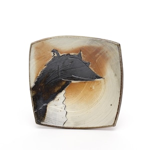 May include: Square ceramic plate with a unique design. The plate features a black abstract shape on a beige and brown background. The plate has a slightly raised edge and a smooth surface. The design is reminiscent of a horse's head.