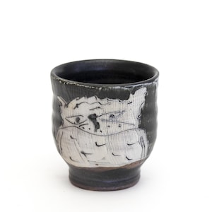Peut inclure: Une tasse en céramique faite à la main avec un extérieur noir et un motif de visage blanc stylisé. La tasse a un intérieur sombre et une surface texturée. La base est de couleur brun foncé.