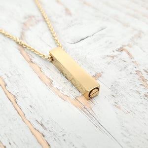 May include: A gold-colored bar pendant necklace. The rectangular pendant has a textured surface and a screw closure at one end. The chain is a delicate gold color, and the necklace is displayed on a white and gray wooden surface.