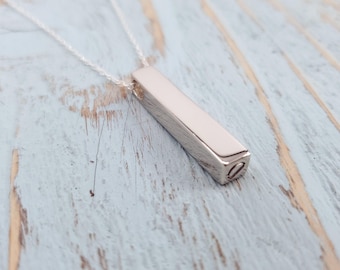 May include: A silver-colored rectangular pendant necklace. The pendant is a small, polished bar suspended from a delicate chain. The necklace is displayed against a light blue and white wooden surface.