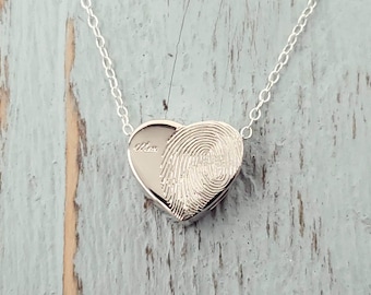 May include: A silver heart-shaped pendant necklace with a fingerprint design on the front and the word "Mom" engraved on the inside. The necklace hangs from a delicate silver chain.