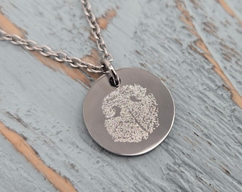 Op de afbeelding: Een zilverkleurige ronde hanger aan een ketting met een gedetailleerde, getextureerde gravure van een hondenneusafdruk op de voorzijde. De hanger is bevestigd aan een bijpassende zilverkleurige metalen ketting met kleine, ovale schakels.