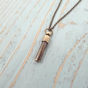 May include: A brass pendant necklace with a long, cylindrical charm. The charm has a rounded top and a smooth, metallic finish.