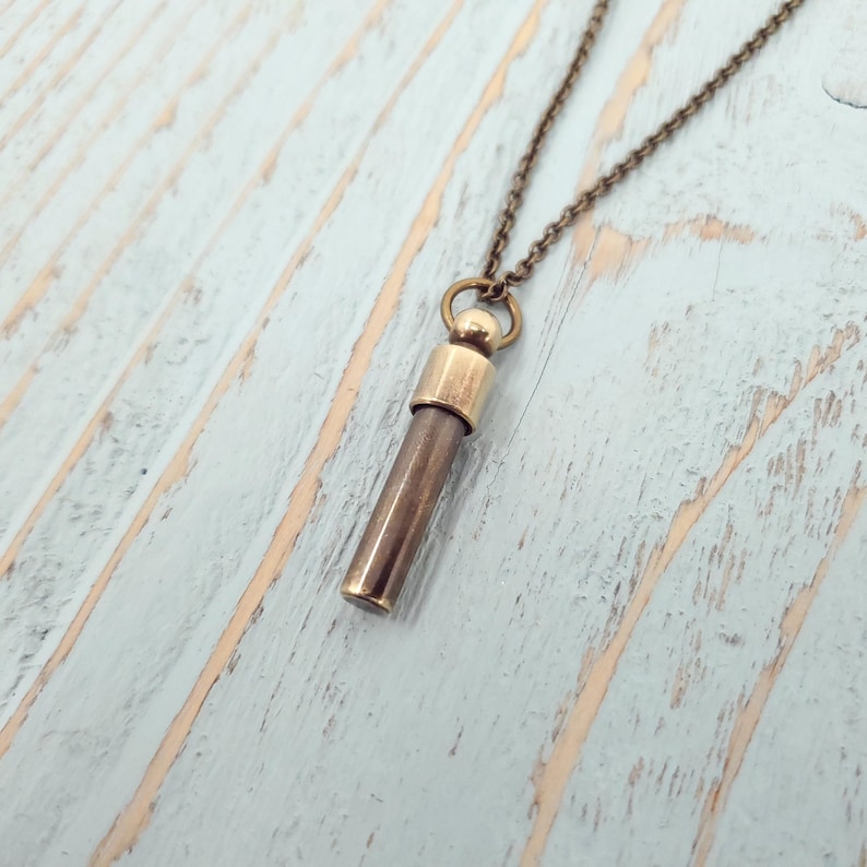 May include: A brass pendant necklace with a long, cylindrical charm. The charm has a rounded top and a smooth, metallic finish.