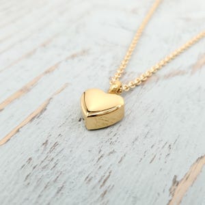 May include: A gold-colored heart-shaped pendant necklace. The pendant is a solid heart with a rounded shape. The chain is delicate and also gold-colored. The necklace is displayed on a light-colored, weathered wooden surface.