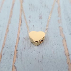May include: A gold-colored heart pendant necklace. The heart-shaped pendant is solid and has a textured surface. The necklace chain is delicate and gold-toned. The background is a light blue and white wooden surface.