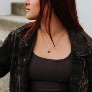 May include: A rose gold necklace with a small star pendant. The necklace is worn over a dark gray ribbed tank top and a black denim jacket. The star pendant is a warm brown color.