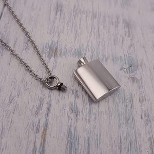 May include: A silver-colored, rectangular pendant necklace with a small flask design. The flask is detached from the chain, revealing the screw-top closure. The chain and flask are made of a shiny, metallic material, resting on a light-colored, textured surface.
