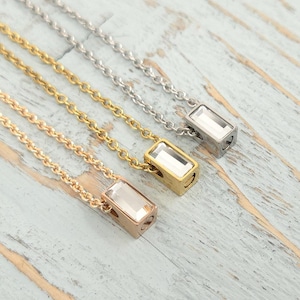 May include: Four necklaces with rectangular crystal pendants. The chains are gold, silver, and rose gold. The pendants are set in matching metal frames. The necklaces are displayed on a light blue and white wooden surface.
