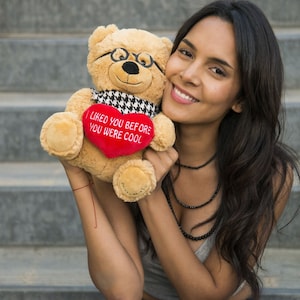 May include: A woman is holding a brown teddy bear with glasses and a red heart that says "I liked you before you were cool".