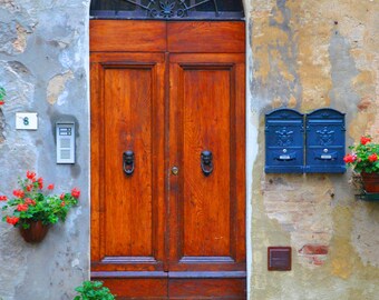 May include: A weathered, light brown wooden door with a black metal grate window above it. The door has two panels and two black metal door handles. The door is set in a stone wall with a number "8" and a doorbell on the left side. There are two blue mailboxes on the right side of the door.
