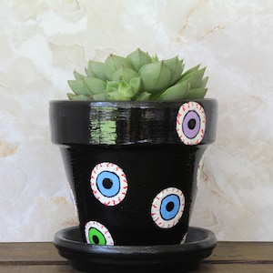 May include: A black painted ceramic flower pot with a matching saucer, decorated with multiple painted eye designs in blue, green, and purple. The pot contains a green succulent plant. The background is a marble-like pattern.