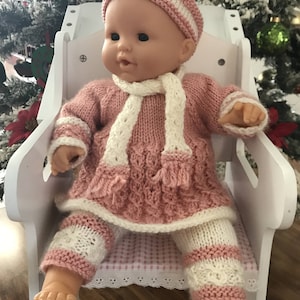May include: A knitted outfit for a doll, in pink with white stripes, comprising a dress, trousers, headband and scarf. The outfit features a delicate cable knit pattern.