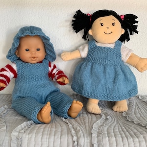 Peut inclure: Deux poupées en tricot portant une salopette bleue et une robe bleue. Les poupées ont les yeux bruns et les joues roses. La poupée de gauche porte une chemise à rayures rouges et blanches. La poupée de droite a les cheveux noirs avec des nœuds roses.