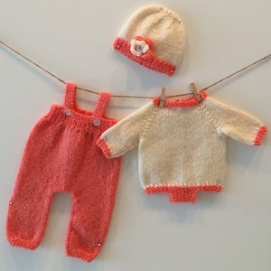 May include: A knitted baby outfit set in pink and white. The set includes a hat, a sweater, and overalls.