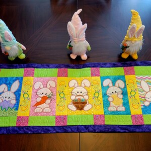 May include: A purple, green, pink, and yellow quilted table runner with five embroidered bunnies. The bunnies are holding carrots, eggs, and baskets. Three plush Easter gnomes with bunny ears are in the background.