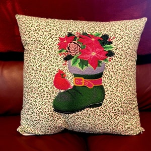 May include: A white pillow with a green boot and red cardinal embroidered on it. The boot has a red and gold buckle and is filled with a bouquet of red poinsettias and a pink flower.