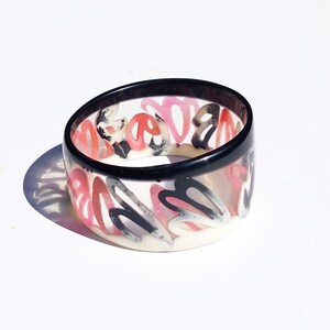 Könnte beinhalten: Ein schwarz-weißes Armreif-Armband mit einem abstrakten Design in Rosa und Weiß. Der Armreif ist aus klarem Kunststoff mit einem schwarzen Rand.