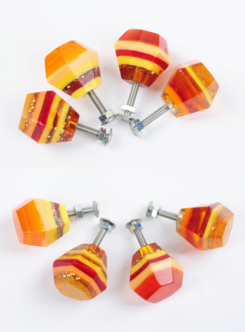 Op de afbeelding: Acht kleurrijke kastknoppen met een geometrische vorm en een gelaagd ontwerp in tinten oranje, geel en rood. De knoppen hebben een zilveren schroef en moer.