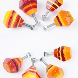 Op de afbeelding: Set van zeven kleurrijke kastknoppen met een geometrische diamantvorm. De knoppen zijn gemaakt van hars en hebben een gelaagd ontwerp met rode, oranje en gele kleuren. Elke knop heeft een zilveren schroef.