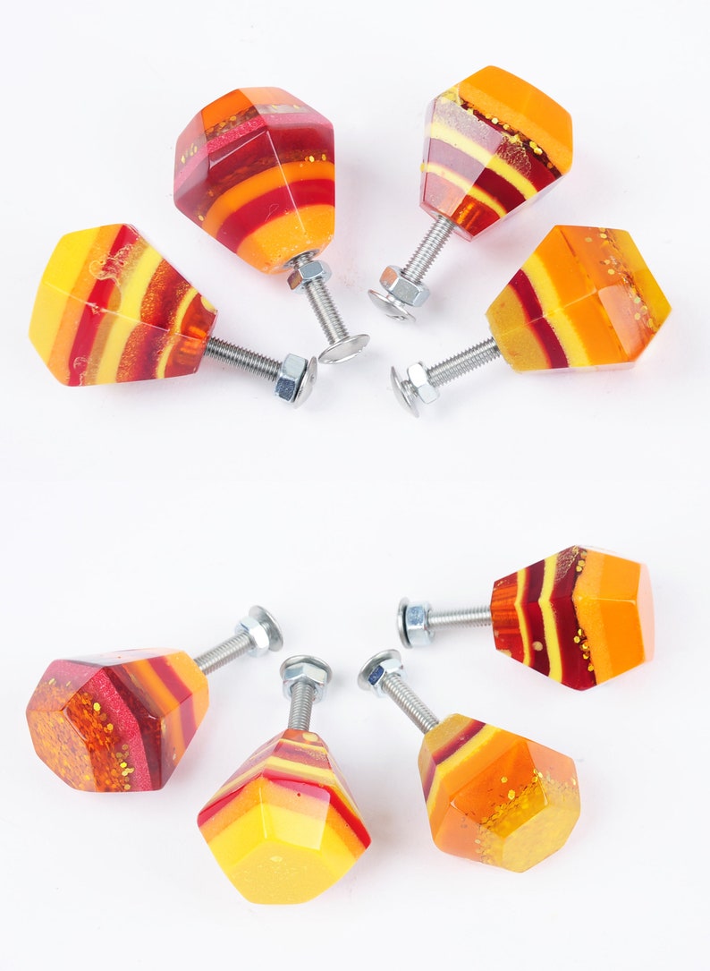 Op de afbeelding: Set van zeven kleurrijke kastknoppen met een geometrische diamantvorm. De knoppen zijn gemaakt van hars en hebben een gelaagd ontwerp met rode, oranje en gele kleuren. Elke knop heeft een zilveren schroef.