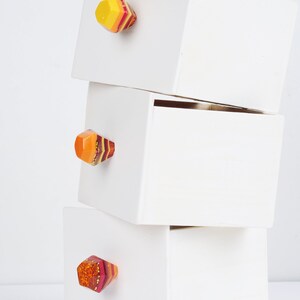 Op de afbeelding: Drie witte houten dozen die op elkaar gestapeld zijn. Elke doos heeft een kleurrijke, geometrische ladegreep met rode, gele en oranje strepen.