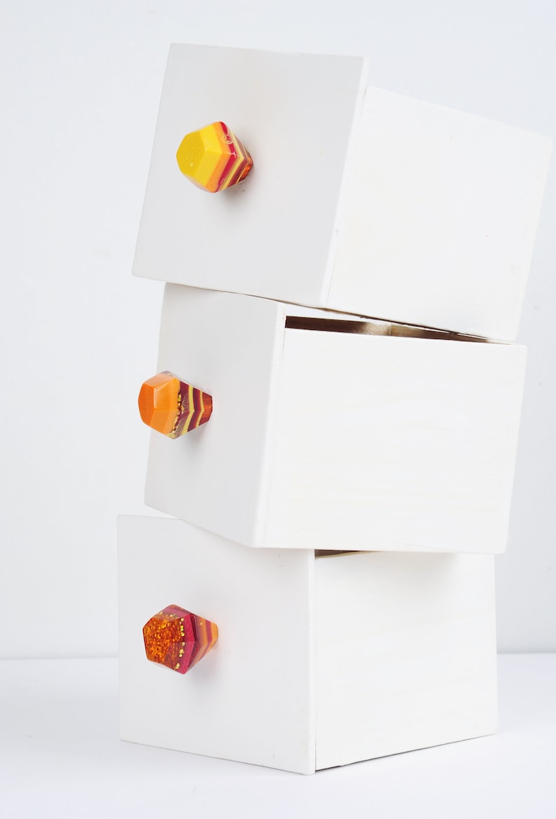 Op de afbeelding: Drie witte houten dozen die op elkaar gestapeld zijn. Elke doos heeft een kleurrijke, geometrische ladegreep met rode, gele en oranje strepen.