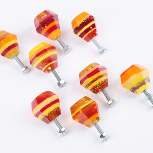 Op de afbeelding: Acht kleurrijke glazen kastknoppen met een geometrische vorm en een zilveren schroef. De knoppen hebben een gelaagd ontwerp met rode, oranje en gele strepen.