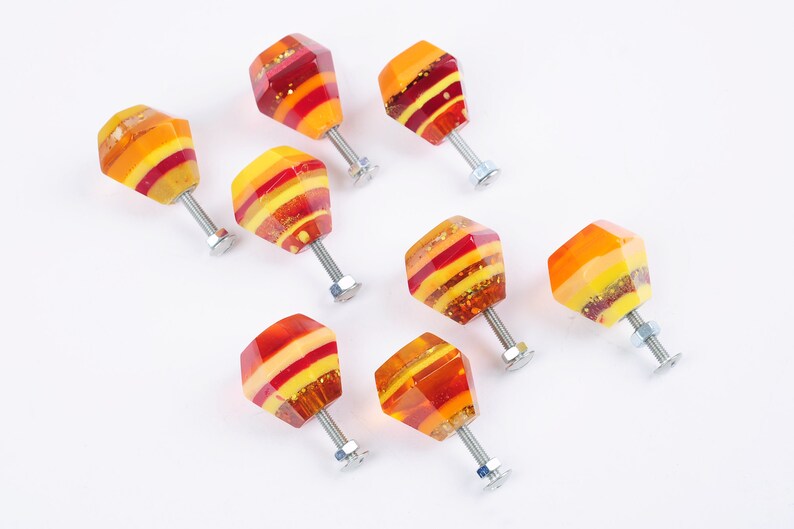 Op de afbeelding: Acht kleurrijke glazen kastknoppen met een geometrische vorm en een zilveren schroef. De knoppen hebben een gelaagd ontwerp met rode, oranje en gele strepen.